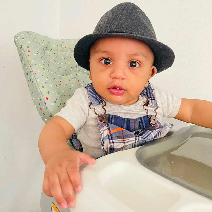 Khristine Spencer joined the competition — help win amazing prizes! baby, child, hat, fedora, high_chair, plaid, overalls, white_wall, cushion, indoor, person, cute, curious, hand, face, seated, infant, clothing, portrait, eyewear