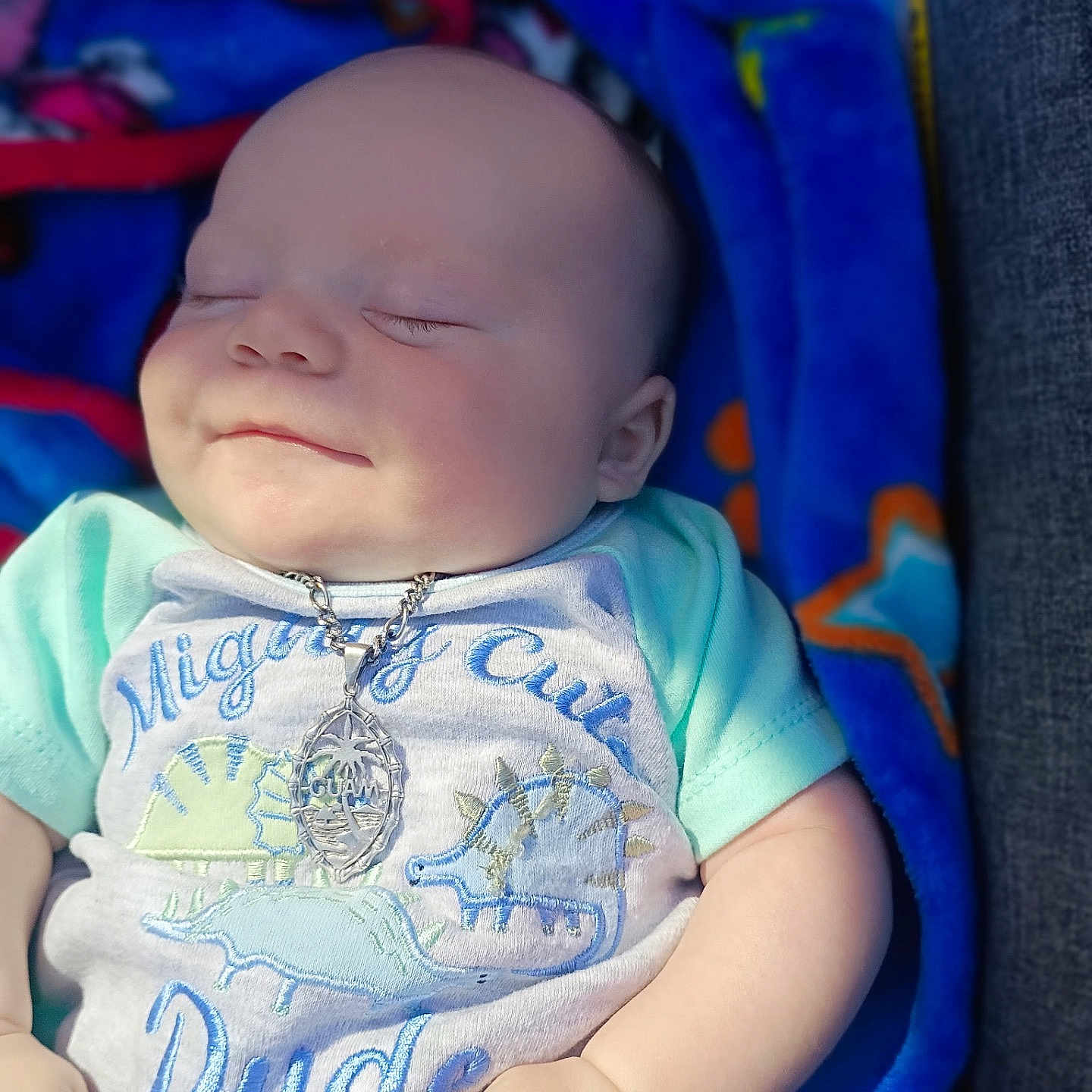 Malakai is registered to the contest to win money with this photo: baby, sleeping, smile, necklace, shirt, dinosaur, blue_blanket, hand, infant, cute, peaceful, child, clothing, portrait, soft_light, indoors, resting, closeup, person, cozy