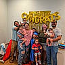 family, children, adults, group_photo, celebration, graduation, congratulations, balloons, gold_balloons, birthday, indoor, portrait, smiling, baby, toddler, school_age, graduation_cap, blue_gown, red_carpet, home