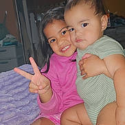 Lance is registered to the contest to win money with this photo: children, siblings, toddler, girl, baby, portrait, smiling, peace_sign, cuddling, sitting, indoor, bedroom, blanket, pink_shirt, striped_clothing, short_hair, long_hair, family, happy, cozy