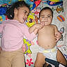 children, siblings, baby, toddler, infant, portrait, smiling, lying_down, bed, blanket, diaper, pink_shirt, long_sleeves, curly_hair, family, indoors, bedroom, cartoon_bedding, holding_hands, candid_photo