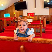 Rosalie is registered to the contest to win money with this photo: baby, child, girl, headband, bow, denim, overalls, pew, church, red_carpet, wood_paneling, smile, happy, interior, furniture, television, cross, candle, religious, cute