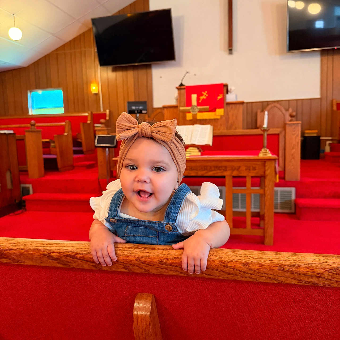 Rosalie is registered to the contest to win money with this photo: baby, child, girl, headband, bow, denim, overalls, pew, church, red_carpet, wood_paneling, smile, happy, interior, furniture, television, cross, candle, religious, cute
