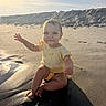 baby, child, beach, sand, sunlight, yellow_clothing, smiling, sitting, waving, water, shadow, outdoor, nature, happy, infant, cute, portrait, sunset, dune, playful