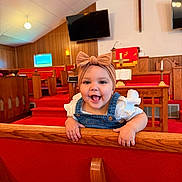 Rosalie is registered to the contest to win money with this photo: baby, child, infant, bow_headband, denim_overalls, wooden_pew, church, cross, red_carpet, wood_paneling, television, candle, smiling, happy, indoor, person, cute, toddler, female, playful