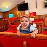 baby, child, infant, bow_headband, denim_overalls, wooden_pew, church, cross, red_carpet, wood_paneling, television, candle, smiling, happy, indoor, person, cute, toddler, female, playful