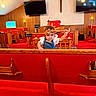 Rosalie joined the competition — help win amazing prizes! toddler, child, church, pew, red_carpet, wooden_furniture, cross, headband, television, candlesticks, red_upholstery, curious, indoor, person, baby, overalls, white_shirt, decor, lighting, playful