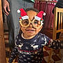 toddler, child, glasses, holiday, christmas, santa_hat, sweater, reindeer, smile, indoors, wooden_chair, floor_tiles, person, happy, festive, cozy, portrait, clothing, cute, home