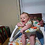 baby, high_chair, smiling, indoor, plant, furniture, child, person, table, chair, window_blinds, picture_frame, colorful, happy, legs, feet, clothing, toy_ring, face, arm