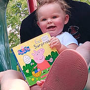 Ava Grace is registered to the contest to win money with this photo: toddler, child, book, peppa_pig, smiling, person, shoe, adidas, outdoor, greenery, tree, leafy, playground, lap, holding, happy, short_sleeve, daylight, footwear, reading