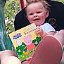 toddler, child, book, peppa_pig, smiling, person, shoe, adidas, outdoor, greenery, tree, leafy, playground, lap, holding, happy, short_sleeve, daylight, footwear, reading