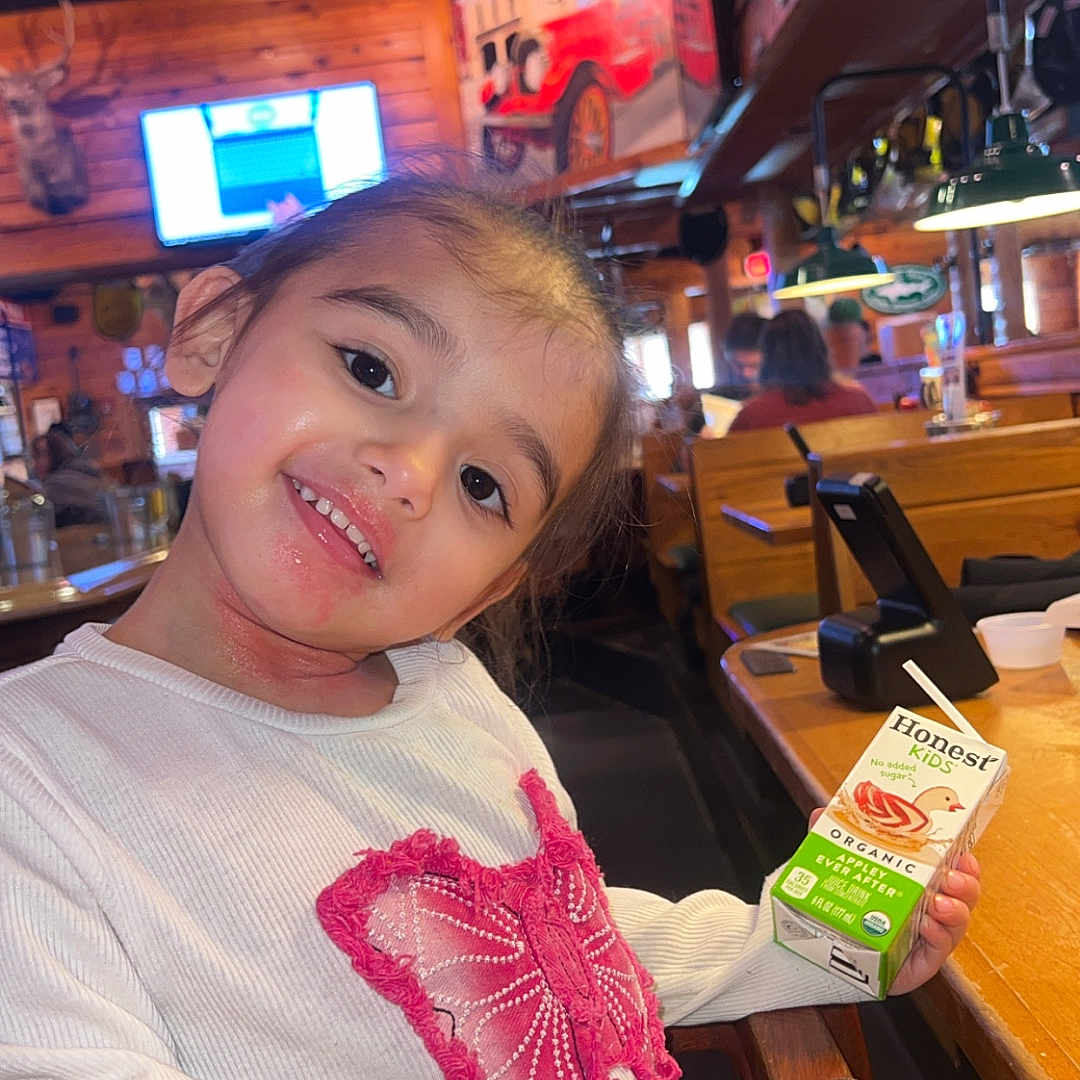 Eliannie is registered to the contest to win money with this photo: casual, child, cozy, happy, indoor, juice_box, lighting, mounted_animal_head, person, pink_butterfly, portrait, restaurant, seated, smiling, table, television, white_sweater, wooden_chair, wooden_interior, young_child