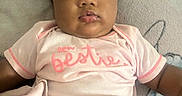 Abrielle is registered to the contest to win money with this photo: baby, infant, child, portrait, lying_down, pink_clothing, onesie, blanket, bed, indoors, face, dark_hair, brown_eyes, serious_expression, looking_at_camera, baby_clothing, text_on_clothing, soft_lighting, cozy, close_up