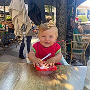 Tom a rejoint le concours — aidez-le/la à gagner de superbes lots ! child, toddler, smiling, red_bib, food_container, spoon, outdoor, cafe, table, sunlight, people, chairs, umbrella, tree, happy, casual, daytime, restaurant, baby, portrait