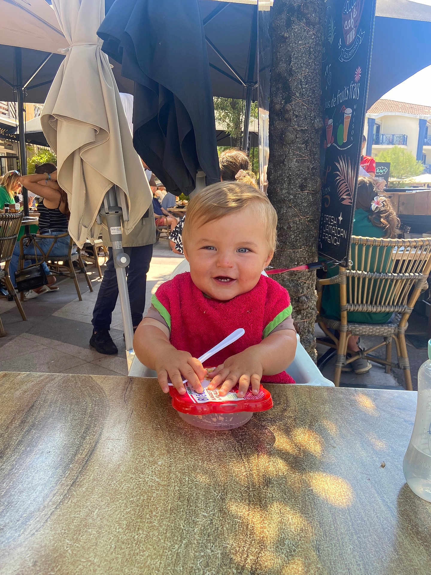 Tom a rejoint le concours — aidez-le/la à gagner de superbes lots ! child, toddler, smiling, red_bib, food_container, spoon, outdoor, cafe, table, sunlight, people, chairs, umbrella, tree, happy, casual, daytime, restaurant, baby, portrait