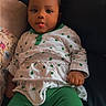 baby, child, infant, person, face, tongue, hair_buns, green_pants, white_top, clover_pattern, sitting, couch, indoor, cute, playful, portrait, young_child, relaxed, smiling, baby_clothing