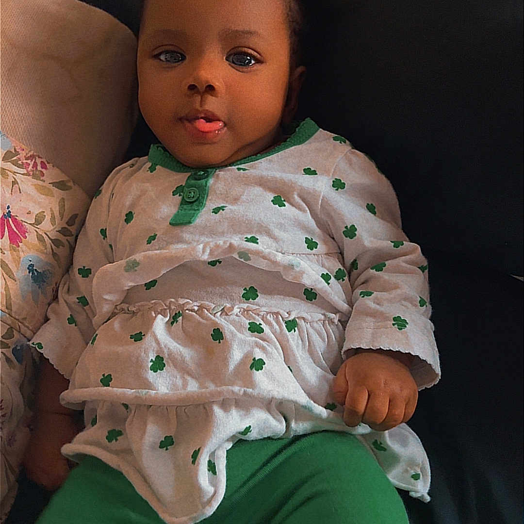 Athena joined the competition — help win amazing prizes! baby, child, infant, person, face, tongue, hair_buns, green_pants, white_top, clover_pattern, sitting, couch, indoor, cute, playful, portrait, young_child, relaxed, smiling, baby_clothing