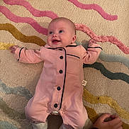 Gracie is registered to the contest to win money with this photo: baby, infant, pink_onesie, carpet, floor, smiling, happy, child, human_hand, pattern, textile, cozy, cute, lying_down, indoor, footwear, sock, buttoned_clothing, face, portrait