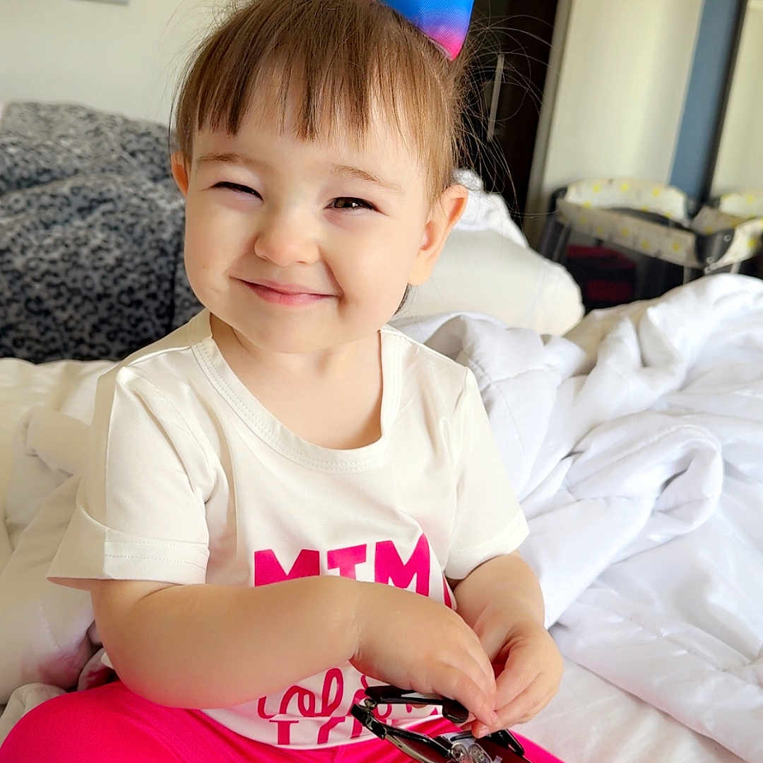 Lila is registered to the contest to win money with this photo: bed, blanket, casual_clothing, child, cute, daylight, furniture, hair_bow, happy, indoor, person, pink_leggings, portrait, rainbow_bow, room, sitting, smiling, sunglasses, toddler, white_shirt