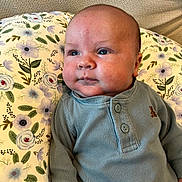 Ezra is registered to the contest to win money with this photo: baby, infant, face, child, blue_shirt, striped_pants, floral_pillow, portrait, closeup, skin, expression, indoor, comfort, soft_light, cute, young, person, head, sitting, cozy