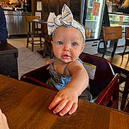 Giada is registered to the contest to win money with this photo: baby, child, headband, bow, blue_eyes, table, wood, high_chair, indoor, restaurant, furniture, person, curious, cute, clothing, face, skin, hand, expression, candid