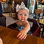 baby, child, headband, bow, blue_eyes, table, wood, high_chair, indoor, restaurant, furniture, person, curious, cute, clothing, face, skin, hand, expression, candid