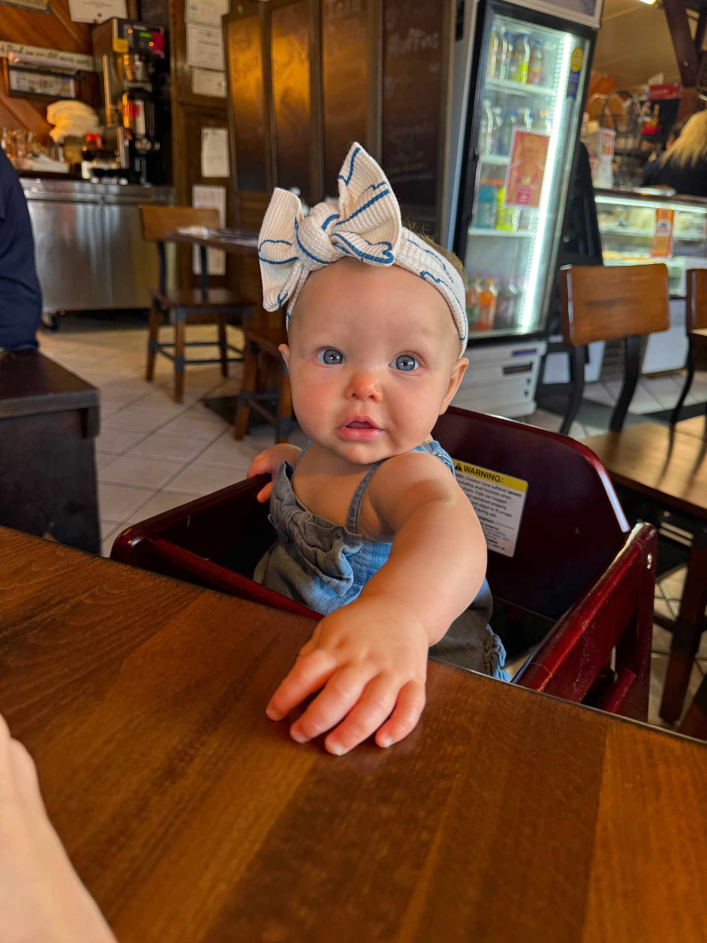 Giada is registered to the contest to win money with this photo: baby, child, headband, bow, blue_eyes, table, wood, high_chair, indoor, restaurant, furniture, person, curious, cute, clothing, face, skin, hand, expression, candid