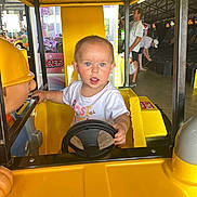 Indikeja joined the competition — help win amazing prizes! adult, amusement, arcade, background, blue_eyes, child, curious, expression, fun, game, indoor, person, ride, seat, short_hair, steering_wheel, toddler, toy, white_shirt, yellow