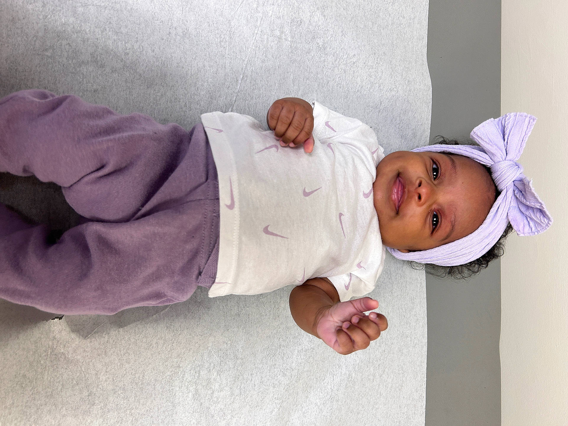 Kehlani is registered to the contest to win money with this photo: baby, bow, child, closeup, clothing, cute, face, hands, happy, headband, headwear, indoor, infant, lying_down, person, portrait, purple_pants, skin, smiling, white_shirt