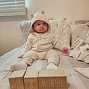 Adriel is registered to the contest to win money with this photo: baby, child, hoodie, bear_ears, wooden_blocks, age_blocks, bed, pillow, blanket, socks, indoors, cute, infant, neutral_expression, soft_lighting, cozy, striped_bedspread, person, portrait, young_child