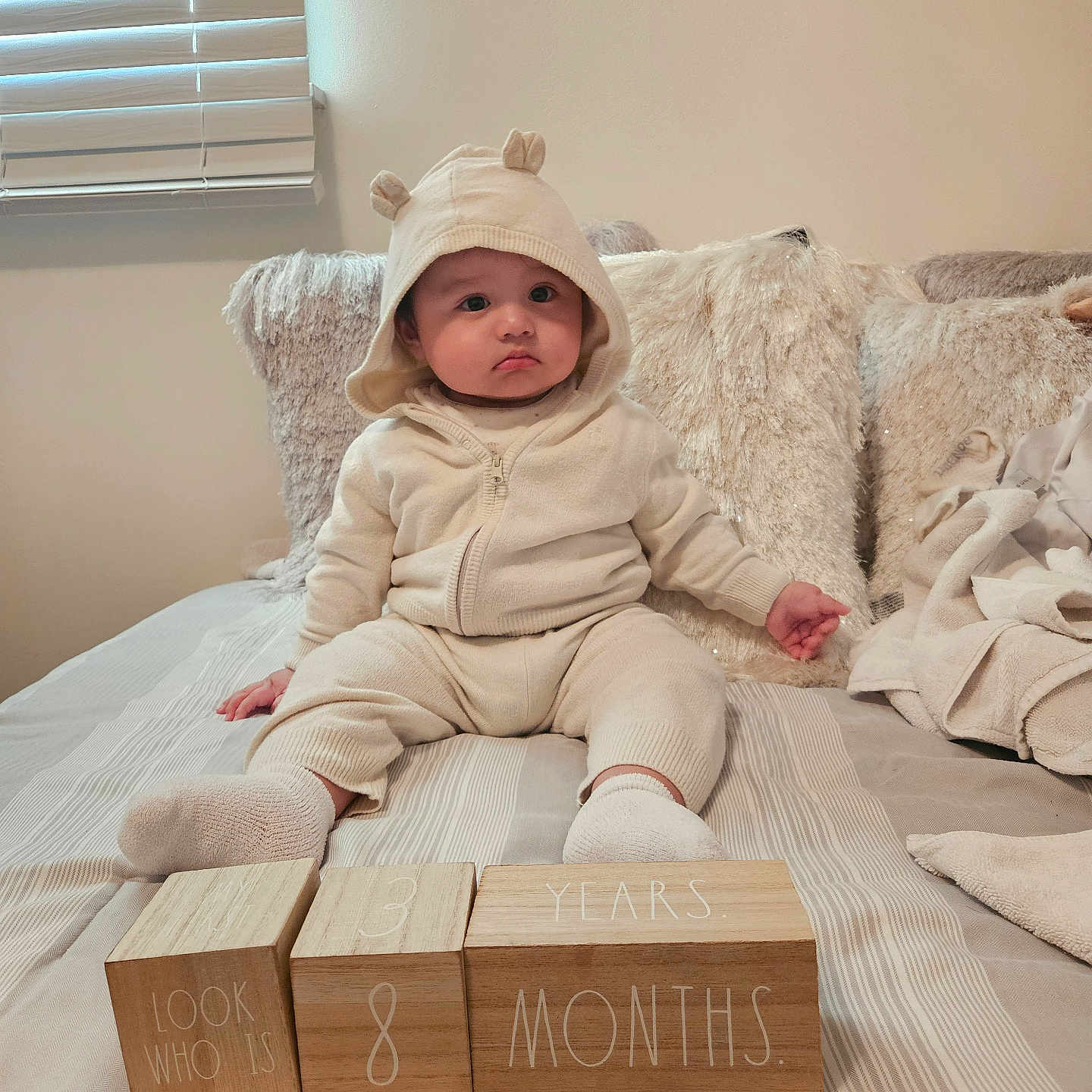 Adriel is registered to the contest to win money with this photo: age_blocks, baby, bear_ears, bed, blanket, child, cozy, cute, hoodie, indoors, infant, neutral_expression, person, pillow, portrait, socks, soft_lighting, striped_bedspread, wooden_blocks, young_child