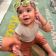 Varun is registered to the contest to win money with this photo: baby, child, sunglasses, smile, hand_holding, sitting, clothing, bracelet, floor_mat, indoor, person, cute, reflection, happy, portrait, infant, casual_wear, accessory, young_child, playful