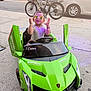 child, toddler, sunglasses, toy_car, green_car, sidewalk, bicycle, parked_cars, street, urban, daylight, open_car_doors, joyful, gesture, reflection, casual_clothing, bracelets, outdoor, playful, vehicle