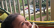 Camilla is registered to the contest to win money with this photo: child, toddler, pacifier, hammock, outdoor, porch, railing, trees, cars, sky, cloudy, curious, colorful, clothing, person, young_child, nature, daytime, relaxing, baby
