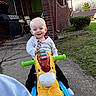 Lillian joined the competition — help win amazing prizes! toddler, child, toy, outdoor, ride_on_toy, plastic_toy, smiling, happy, concrete_path, brick_house, green_lawn, bushes, daylight, casual_clothing, person, play, handlebars, grass, window, sidewalk