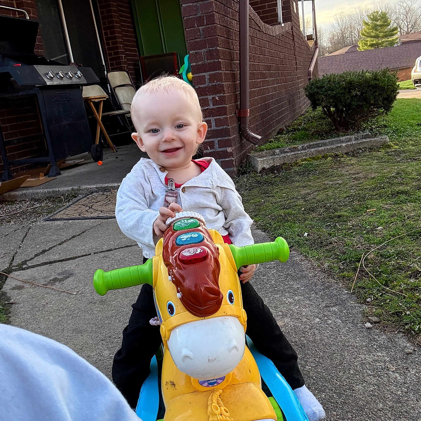 Lillian joined the competition — help win amazing prizes! toddler, child, toy, outdoor, ride_on_toy, plastic_toy, smiling, happy, concrete_path, brick_house, green_lawn, bushes, daylight, casual_clothing, person, play, handlebars, grass, window, sidewalk