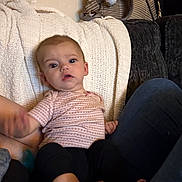 Paislyn is registered to the contest to win money with this photo: baby, blanket, couch, striped_bag, patterned_shirt, black_pants, indoor, person, child, seated, cozy, furniture, legs, comfort, curious, young_child, soft_lighting, home_interior, relaxed, textile