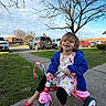 toddler, tricycle, outdoor, child, smiling, barefoot, blue_jacket, fruit_pattern_dress, sidewalk, grass, tree, sky, clouds, cars, parking_lot, playful, happy, person, daylight, residential_area