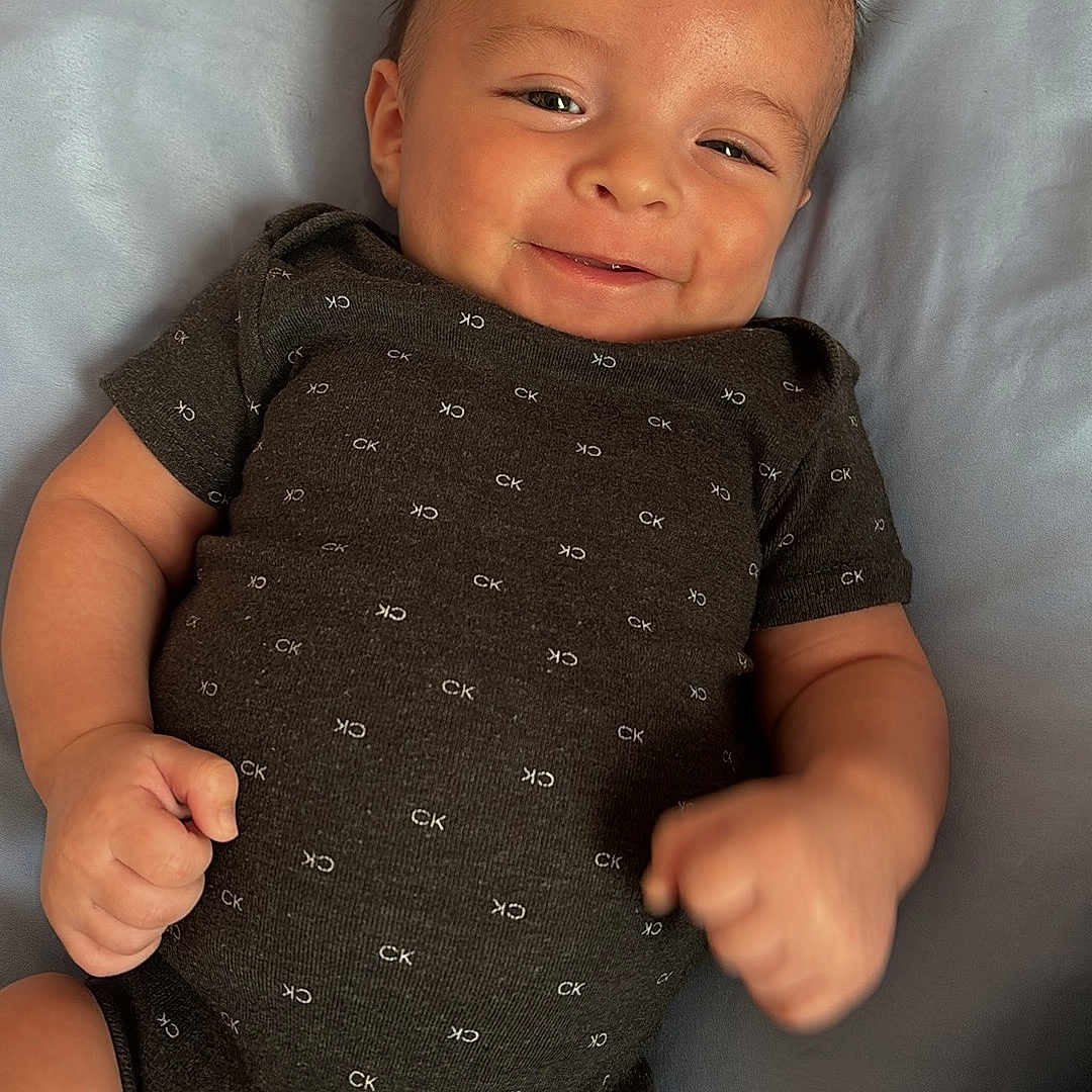 Emilio Ray joined the competition — help win amazing prizes! baby, cheeks, child, clothing, cute, fabric, face, fist, happy, head, indoors, infant, lying_down, onesie, pattern, short_sleeves, skin, smile, soft_surface, young