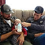 adult, baby, beard, blue_jeans, bottle, cap, care, casual_clothing, couch, curtains, family, feeding, floral_pattern, hand, hat, indoor, long_sleeve, man, plaid_shirt, red_sleeves