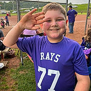 Davis is registered to the contest to win money with this photo: child, boy, smiling, purple_shirt, sports_jersey, number_77, baseball_field, chain_link_fence, sunset, outdoor, grass, spectators, people, hat, water_bottle, team, recreation, fun, game, happy