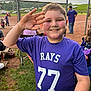 child, boy, smiling, purple_shirt, sports_jersey, number_77, baseball_field, chain_link_fence, sunset, outdoor, grass, spectators, people, hat, water_bottle, team, recreation, fun, game, happy
