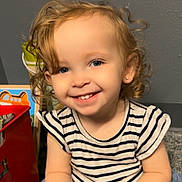 Layla is registered to the contest to win money with this photo: toddler, child, smile, curly_hair, striped_shirt, toy, indoor, happy, portrait, person, blue_eyes, earring, playful, cute, sitting, young_child, casual_clothing, small_toy, light_skin, background_objects