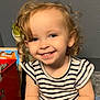 toddler, child, smile, curly_hair, striped_shirt, toy, indoor, happy, portrait, person, blue_eyes, earring, playful, cute, sitting, young_child, casual_clothing, small_toy, light_skin, background_objects