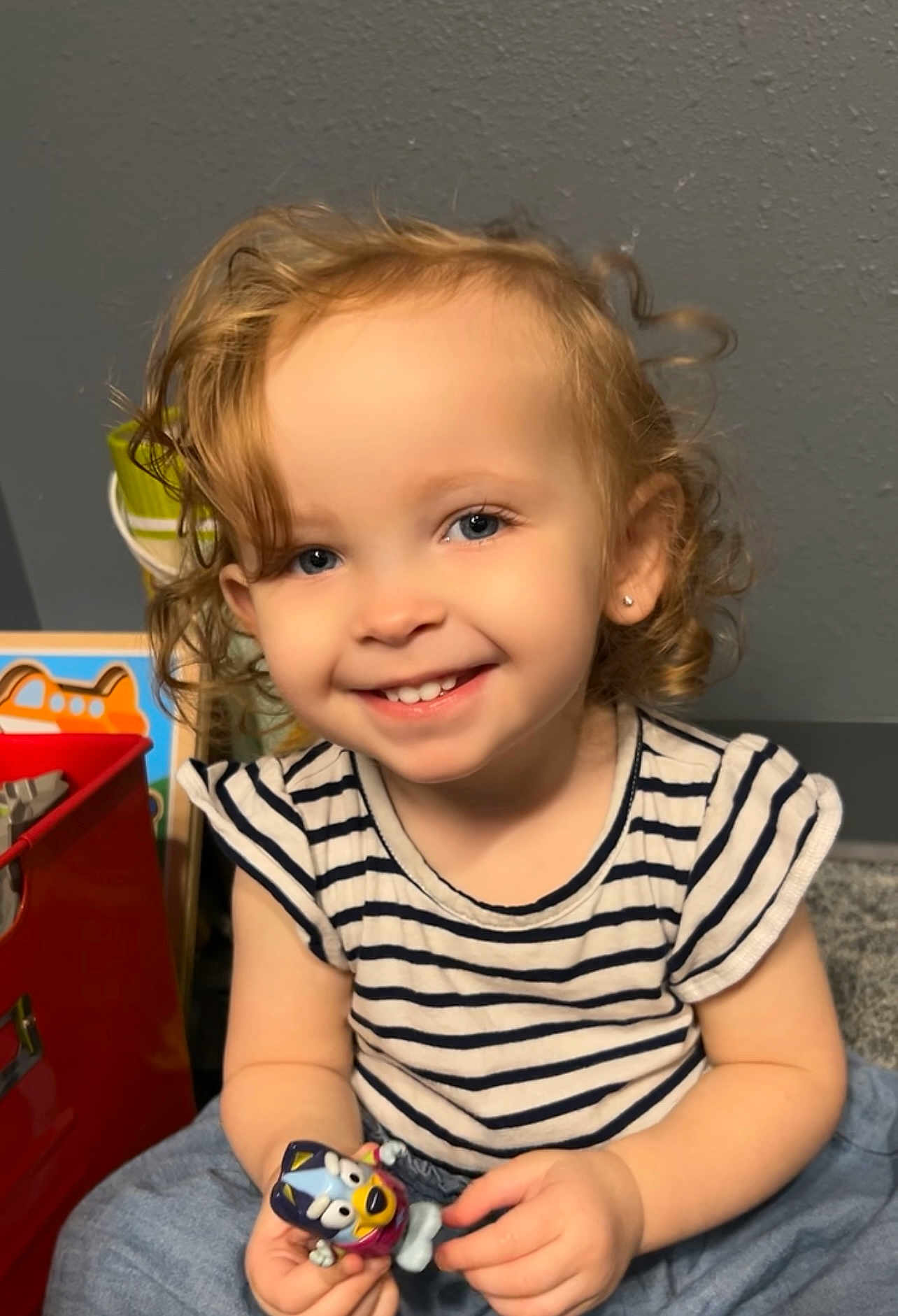 Layla is registered to the contest to win money with this photo: toddler, child, smile, curly_hair, striped_shirt, toy, indoor, happy, portrait, person, blue_eyes, earring, playful, cute, sitting, young_child, casual_clothing, small_toy, light_skin, background_objects