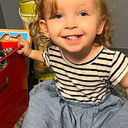 Layla is registered to the contest to win money with this photo: blue_skirt, carpet, child, colorful, container, curly_hair, cute, earrings, floor, happy, indoor, person, playtime, portrait, smiling, striped_shirt, toddler, toy, wall, young_child