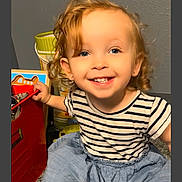 Layla is registered to the contest to win money with this photo: blue_eyes, blue_skirt, carpet, casual_clothing, child, curly_hair, earrings, floor, happy, indoor, person, playful, portrait, room, smiling, striped_shirt, toddler, toy_chest, toys, young_child