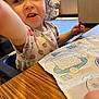 Layla is registered to the contest to win money with this photo: activity_sheet, background, blue_eyes, child, crayon, curly_hair, daylight, expression, face, floral_pattern, hand, highchair, indoor, maze, seated, shirt, table, toddler, window, wooden_table