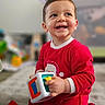 toddler, child, smiling, red_onesie, toy, indoor, carpet, playing, happy, baby, cute, kneeling, young_child, colorful_toy, face, person, home, floor, expression, clothing