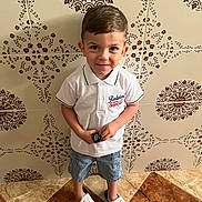 Iyad participe au concours pour gagner de l'argent avec cette photo : child, boy, standing, white_polo, denim_shorts, nike_sandals, tile_floor, patterned_wall, smiling, short_hair, indoor, casual_clothing, holding_object, footwear, portrait, happy, cute, playful, young, person